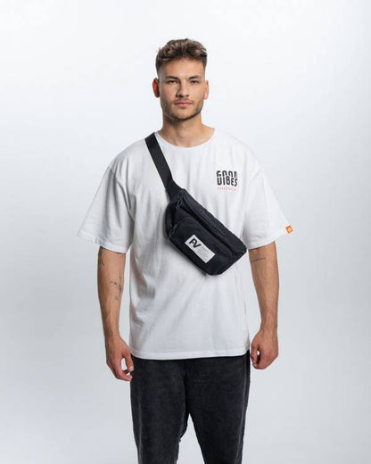 (DE) Ein Mann steht vor einem weißen Hintergrund. Er trägt ein weißes T-Shirt mit dem dicken schwarzen  Schriftzug "GOOD VIBES" und dazu eine Bauchtasche. (EN) A man stands in front of a white background. He is wearing a white T-shirt with the thick black lettering ‘GOOD VIBES’ and a fanny pack.