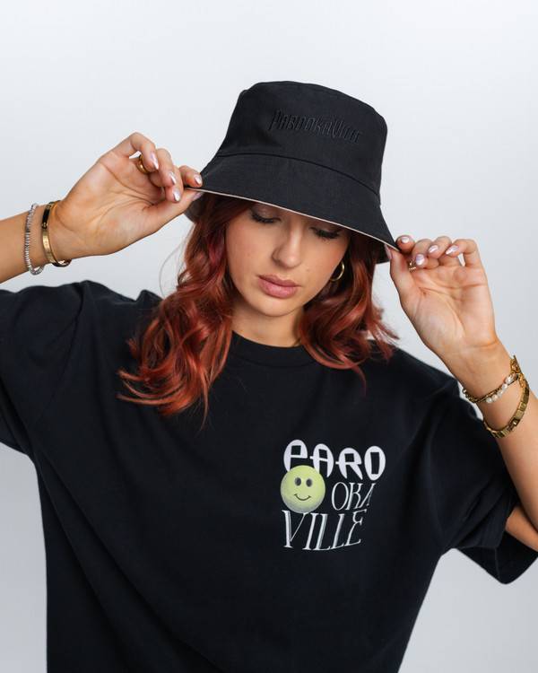 (DE) Eine Frau trägt ein schwarzes T-Shirt mit weißer PV Schrift.  Außerdem ist links im Schriftzug ein grüner Smiley. Dazu trägt sie einen schwarzen Bucket Hat . (EN) A woman is wearing a black T-shirt with white PV lettering.  There is also a green smiley face in the lettering on the left. She is also wearing a black bucket hat.