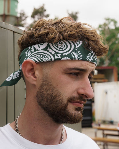 (DE) Ein Mann trägt eine dunkelgrüne Bandana it weißem Blumenmuster um seine Stirn gewickelt. (EN) A man wears a dark green bandana with a white floral pattern wrapped around his forehead.