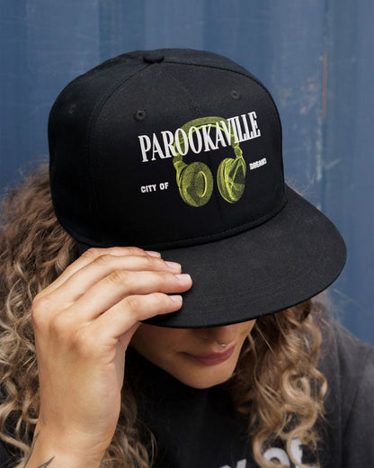 (DE) Eine Frau trägt ein dunkles T-Shirt mit weißer Aufschrift . Dazu trägt sie eine schwarze Cap mit der großen, weißen Aufschrift "PAROOKAVILLE" und der kleineren Unterschrift "CITY OF DREAMS" und einem Image von grünen headphones.  (EN) A woman is wearing a dark T-shirt with white lettering. She is also wearing a black cap with the large, white inscription ‘PAROOKAVILLE’ and the smaller signature ‘CITY OF DREAMS’ and an image of green headphones.