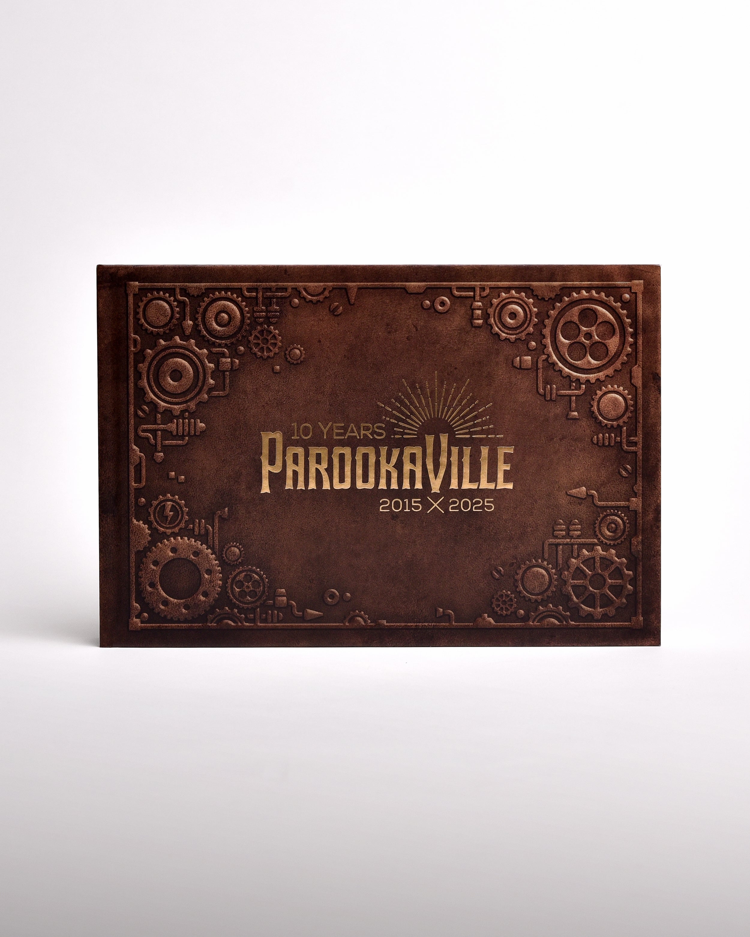 Zu sehen ist ein exklusives Buch im senkrecht-länglichen Format, frontal fotografiert. Es hat einen Steampunk-Artigen Einbund und 3D Förmige Zahnräder an den Rändern gedruckt. Mittig ist das "10 Years Parookaville" Logo in gold-schimmernder Farbe gedruckt.
