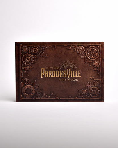 Zu sehen ist ein exklusives Buch im senkrecht-länglichen Format, frontal fotografiert. Es hat einen Steampunk-Artigen Einbund und 3D Förmige Zahnräder an den Rändern gedruckt. Mittig ist das "10 Years Parookaville" Logo in gold-schimmernder Farbe gedruckt.