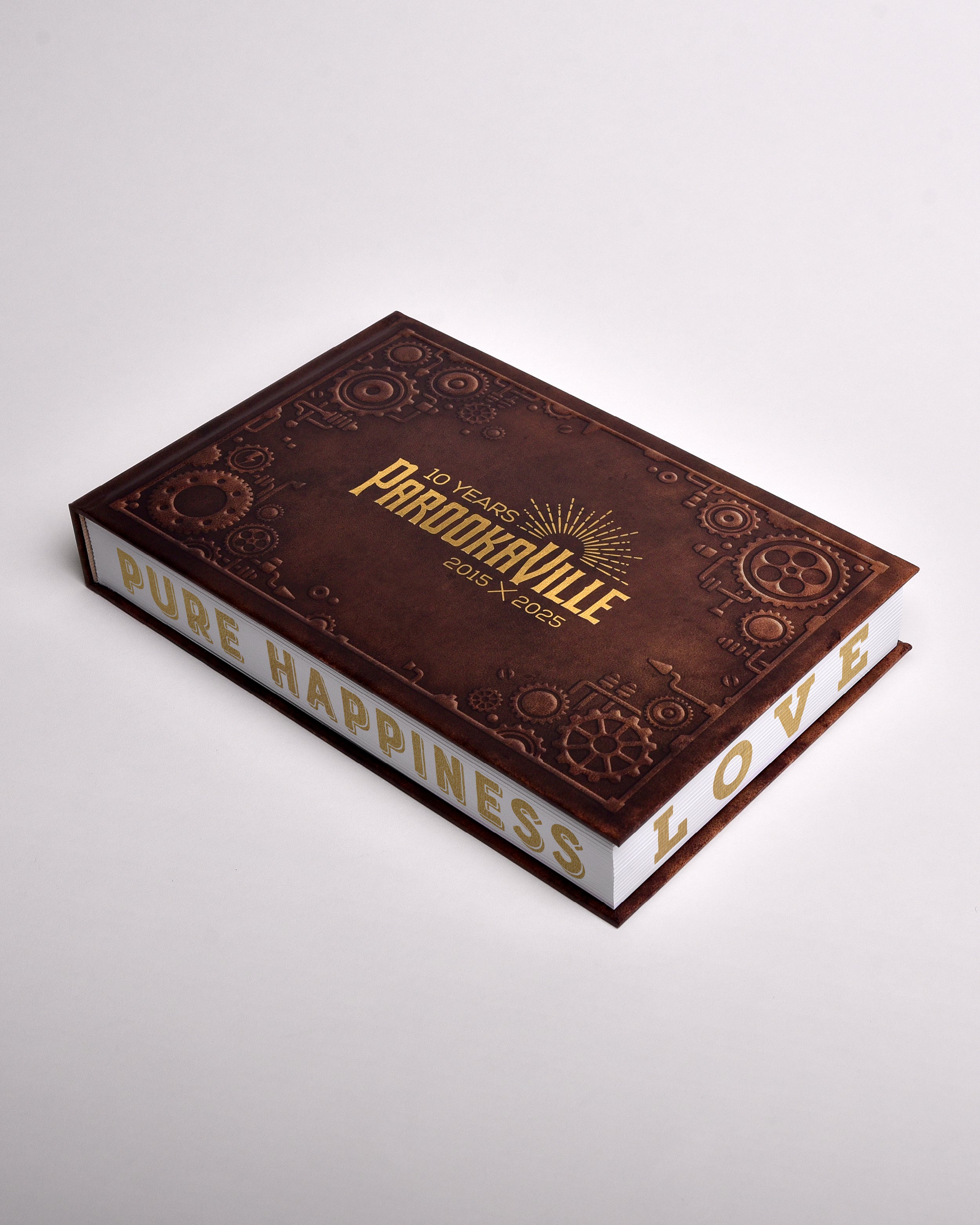Zu sehen ist ein exklusives Buch im sekrecht-länglichen Format. Es hat einen Steampunk-Artigen Einbund und 3D Förmige Zahnräder an den Rändern gedruckt. Mittig ist das "10 Years Parookaville" Logo in gold-schimmernder Farbe gedruckt. Die weißen Seiten sind von außen bedruckt. Unten ist "Pure Happiness" und seitlich "Love" zu lesen.