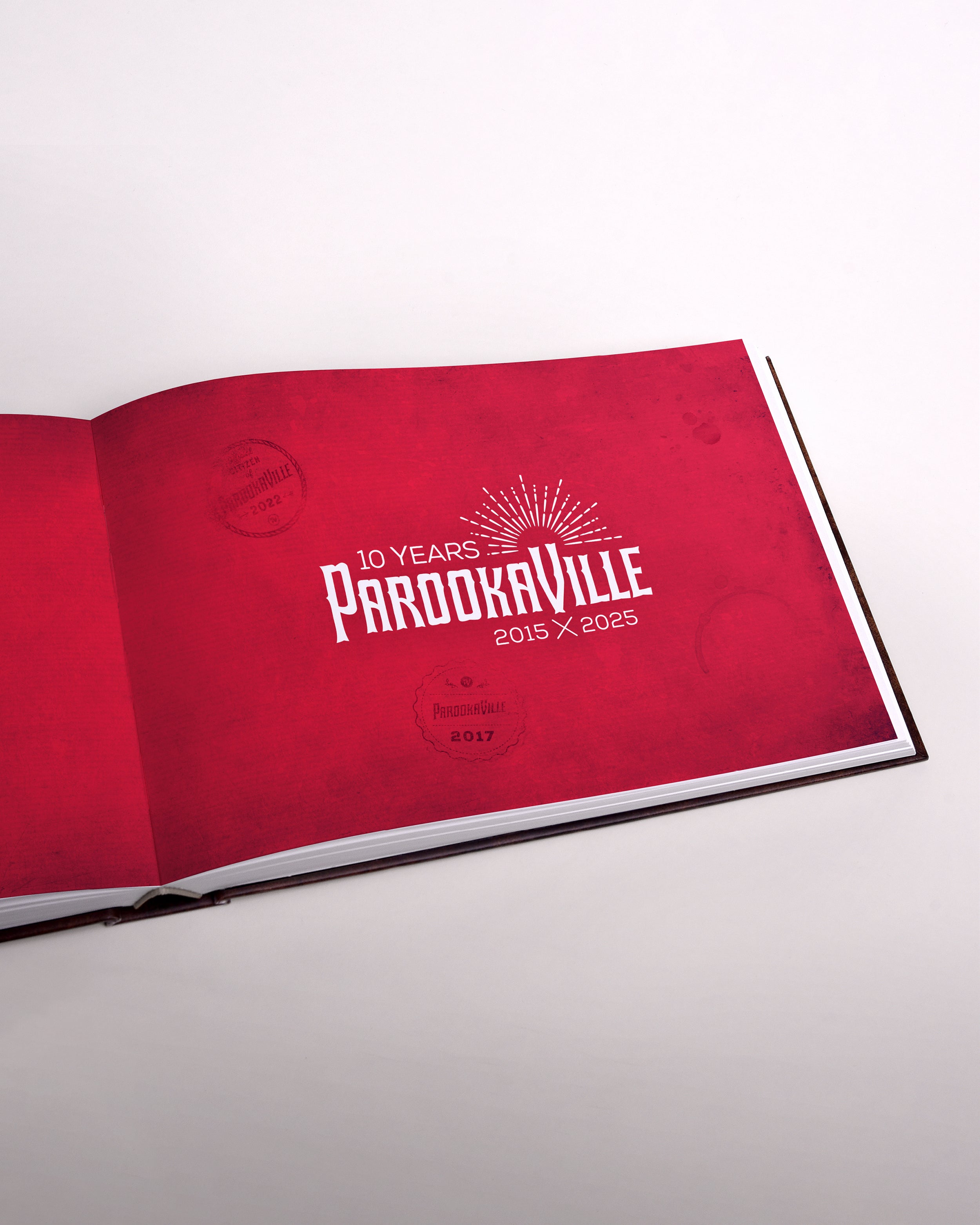 Zu sehen ist eine Seite des Buches im senkrecht-länglichen Format. Die Seite ist rot mit dem weißen "10 Years Parookville" Logo in der Mitte. Es sind leichte Abdrücke der City-Stempel verteilt zu sehen.