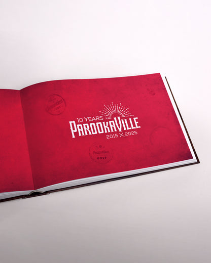 Zu sehen ist eine Seite des Buches im senkrecht-länglichen Format. Die Seite ist rot mit dem weißen "10 Years Parookville" Logo in der Mitte. Es sind leichte Abdrücke der City-Stempel verteilt zu sehen.