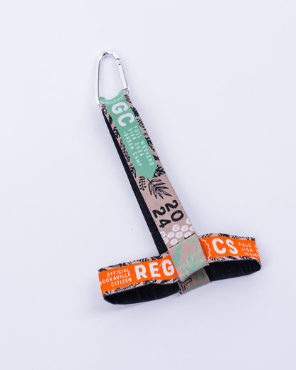 Ein Cup Holder aus upgecycelten Parookaville-Festivalbändern liegt flach auf hellem Hintergrund. Ein silberfarbener Karabiner dient zur Befestigung. Der Tragegurt und der Becherhalter bestehen aus verschiedenfarbigen Festivalbändern mit unterschiedlichen Mustern und Aufdrucken.