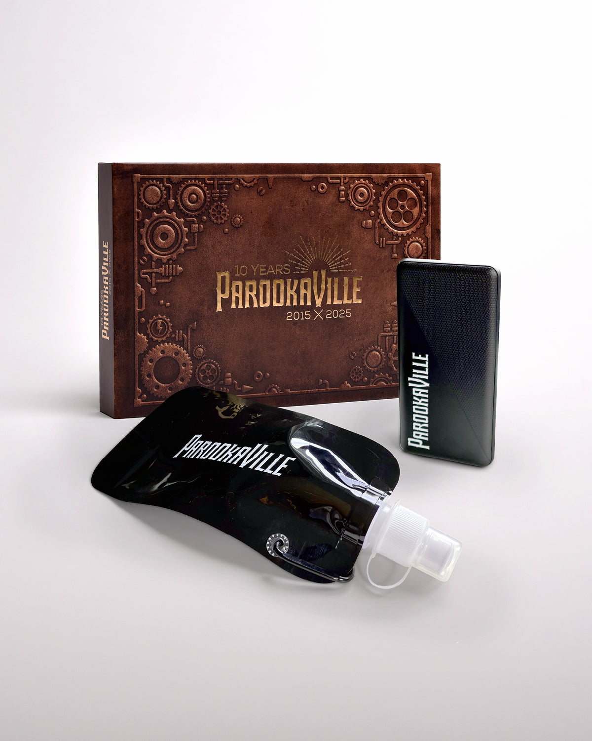 (DE) Drei Artikel auf weißem Hintergrund: das ‚10 Jahre‘-Buch mit sichtbarem Cover, eine schwarze Powerbank mit Parookaville-Logo sowie eine schwarze Faltflasche mit Logo, die davor platziert ist.
(ENG) Three items on a white background: the ‘10 Years’ book with the cover visible, a black power bank featuring the Parookaville logo, and a black collapsible bottle with logo placed in front.