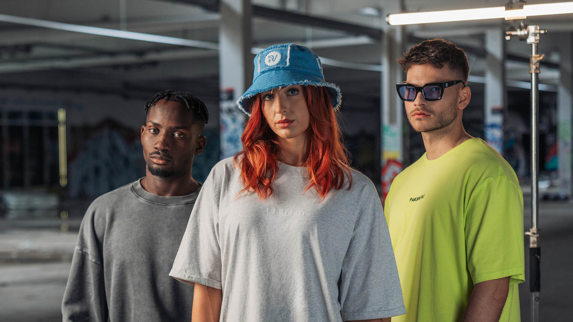 (DE) Drei Personen stehen vor einem bunten Graffiti Hintergrund. Der Mann rechts trägt einen grauen Pullover, die Frau in der Mitte ein graues Oversized T-Shirt und  der Man rechts trägt ein neongrünes T-Shirt. (EN) Three people are standing in front of a colourful graffiti background. The man on the left is wearing a grey sweater, the woman in the middle a light grey T-dress, and the man on the right a neon green T-shirt.