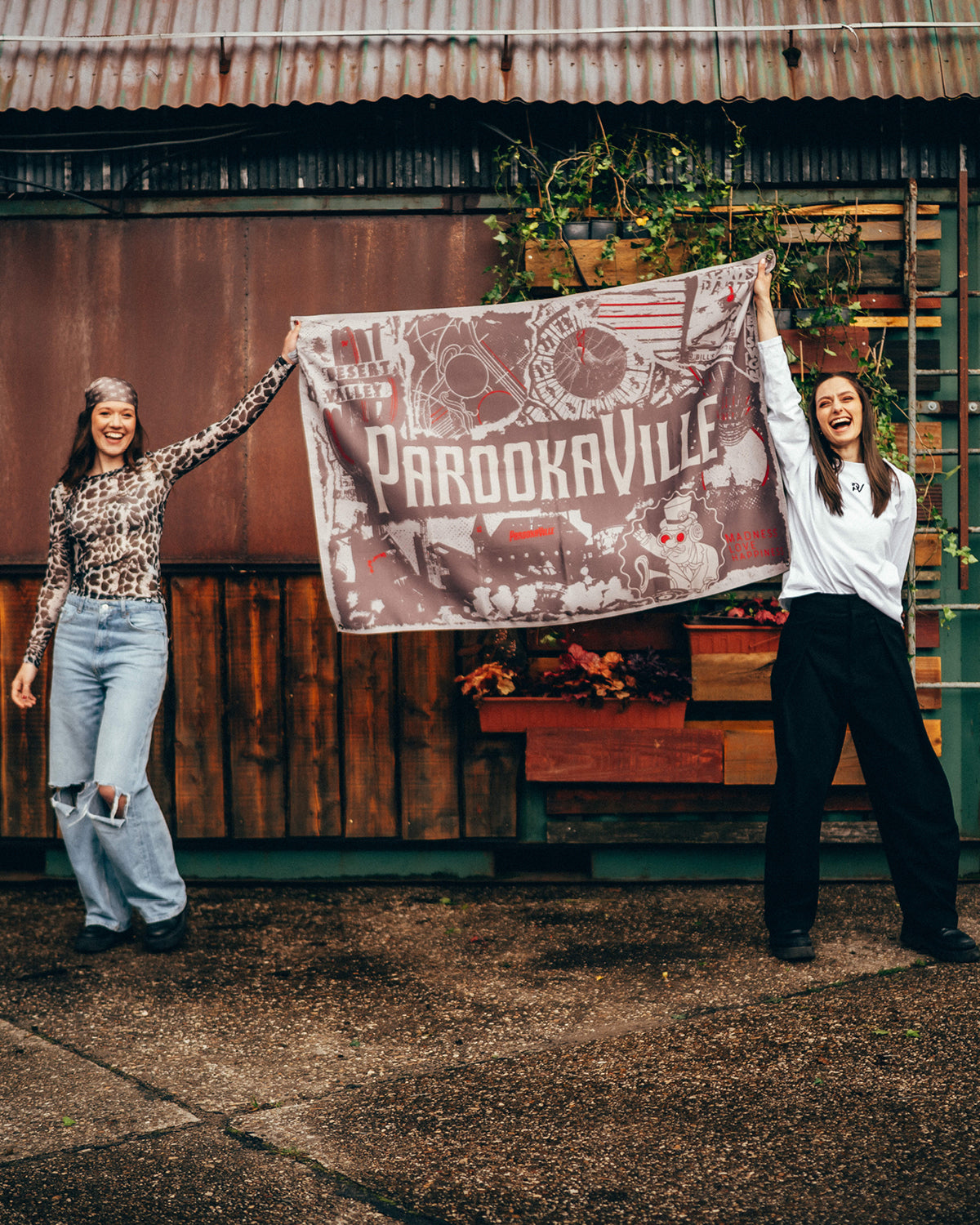 Zwei lachende Frauen halten die große City-Poster-Flagge in warmen Erd- und Beigetönen vor einem Festivalstand mit Holz-, Metall- und Pflanzenelementen hoch. Der Print zeigt den weißen Parookaville-Schriftzug auf einer Collage mit Desert Valley, Kompass-Motiv, Bill Parooka und weiteren grafischen Elementen. 