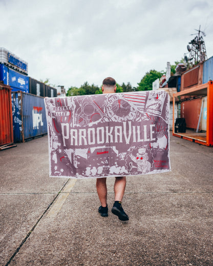 Ein Mann trägt die City-Poster-Flagge in warmen Erd- und Beigetönen mit roten Akzenten ausgebreitet über den Schultern und läuft durch ein Container-Gelände. Von hinten sind das Collagen-Design mit Parookaville-Schriftzug, Desert Valley, Bill Parooka und weiteren grafischen Elementen zu sehen.