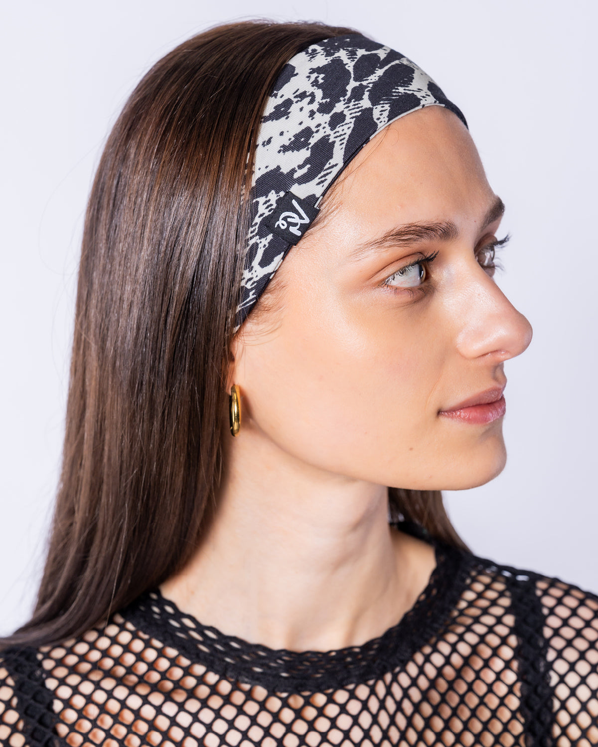 Eine Frau im Seitenprofil trägt das Leo-Haarband mit einem kleinen schwarzen PV-Monogramm-Label an der Seite. Das schwarz-weiße Animal-Print-Band wird zu einem schwarzen Netzoberteil kombiniert.