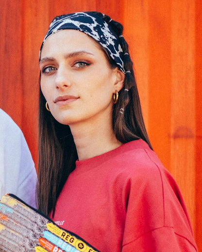 Eine Frau vor einer orangefarbenen Containerwand trägt das schwarz-weiße Leo-Haarband über der Stirn. Das Animal-Print-Accessoire wird zu einem roten T-Shirt und bunten Festivalbändchen an einer kleinen Tasche kombiniert.