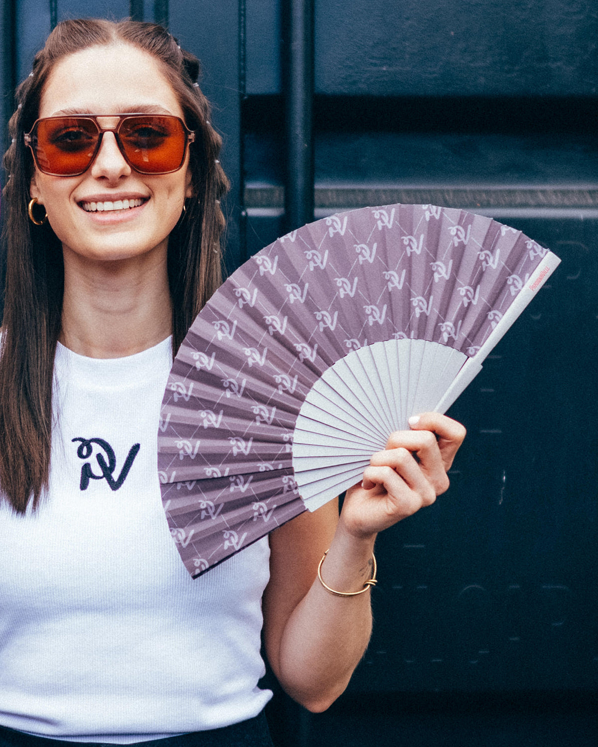 Eine lächelnde Frau mit brauner Sonnenbrille hält den PV-Signature-Handfächer in der Hand. Die Bespannung in dunklem Taupe zeigt ein Muster aus beigefarbenen PV-Monogrammen und feinen Linien. Der Handfächer mit grauem Gestell wird vor einer dunkelblauen Containerwand präsentiert.