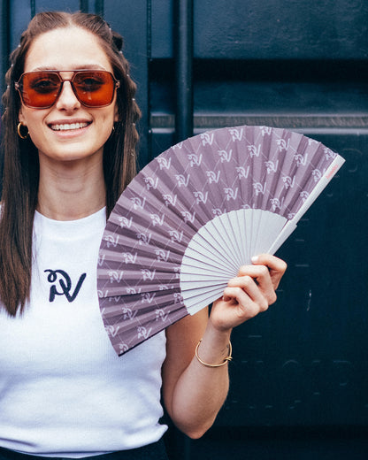 Eine lächelnde Frau mit brauner Sonnenbrille hält den PV-Signature-Handfächer in der Hand. Die Bespannung in dunklem Taupe zeigt ein Muster aus beigefarbenen PV-Monogrammen und feinen Linien. Der Handfächer mit grauem Gestell wird vor einer dunkelblauen Containerwand präsentiert.