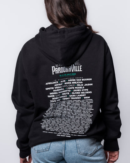 (DE) Eine Frau trägt den schwarzen Line-Up Hoodie mit weißer Schrift auf dem Back Print. Auf der Rückseite stehen die Artists unter der 10 Years PAROOKAVILLE Aufschrift. (EN) A woman wears the black line-up hoodie with white lettering on the back print. On the back are the artists under the 10 Years PAROOKAVILLE inscription.