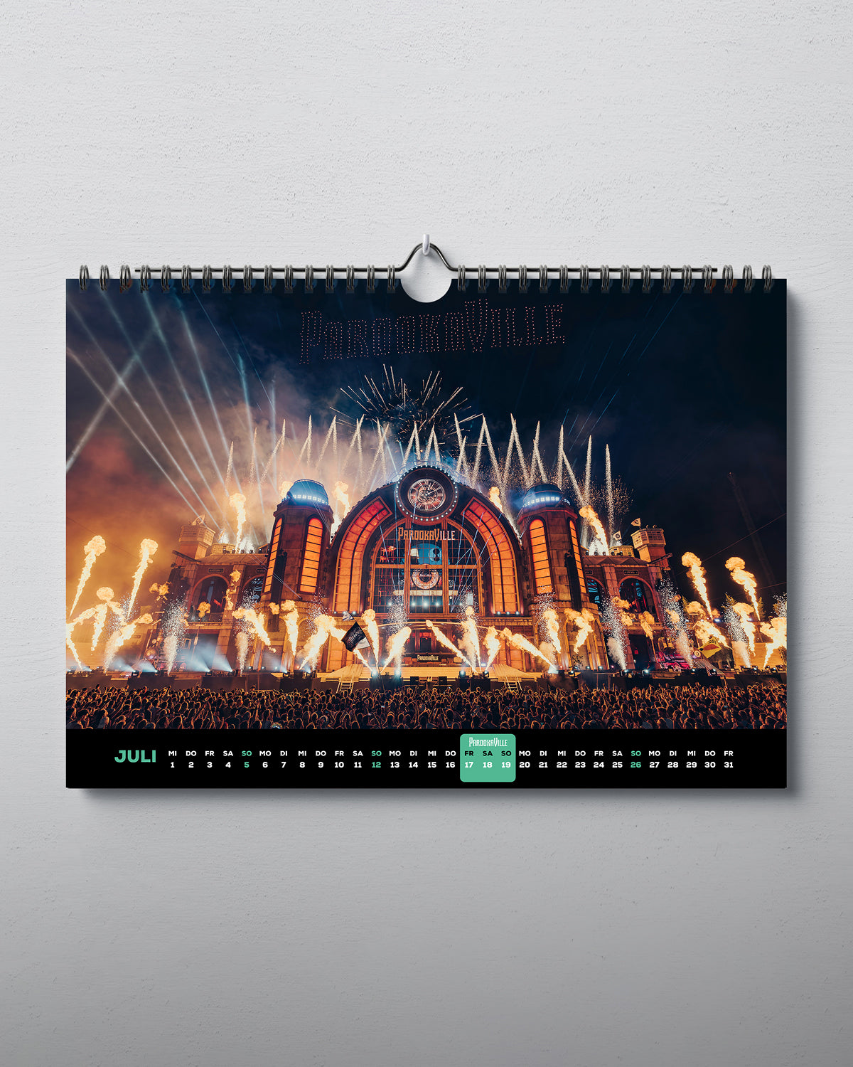 Zu sehen ist der Juli des horizontal aufhängbaren Kalenders. Mittig abgedruckt ist eine Aufnahme der Drohnenshow der Mainstage aus 2025. Unter dem Bild ist ein Streifen mit den Tagen und Wochenenden des Monats. Die Veranstaltungstage für 2026 sind farblich hinterlegt.