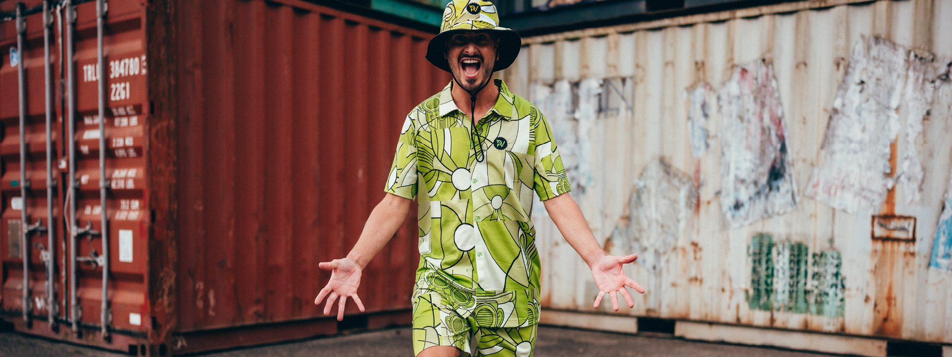 Ein braunhaariger Mann hat das Hemd, die Shorts und den Safari Hut aus der Merchandise Kollektion 2025 im grünen Allover Bill-Print an. Er steht vor Containern, streckt aufgeregt die Arme aus und macht einen Schritt nach Vorne, Die Mimik mit offenen Mund und großen Augen unterstreicht seine positive Energie.