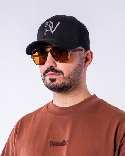 Ein Mann trägt die Urban Aviator Sonnenbrille mit eckigen, braun getönten Gläsern und einem transparent braunen Rahmen. Die Nahaufnahme zeigt das markante Design der Brille, kombiniert mit einer schwarzen Cap mit hellgrauem PV-Logo vor hellem Hintergrund.