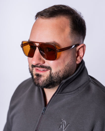 Ein Mann trägt die Urban Aviator Sonnenbrille mit eckigen, braun getönten Gläsern, einem transparent braunen Rahmen und markantem Doppelsteg. Die Nahaufnahme hebt das moderne Aviator-Design und die klaren Linien der Brille vor hellem Hintergrund hervor.