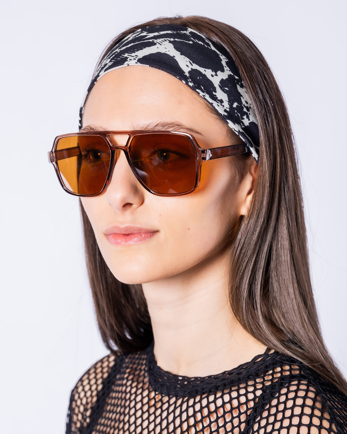 Eine Frau trägt die Urban Aviator Sonnenbrille mit eckigen, braun getönten Gläsern, transparent braunem Rahmen und markantem Doppelsteg. Kombiniert mit einem schwarz-weißen Leo-Stirnband und einem schwarzen Netzoberteil wird die Brille in einer Nahaufnahme vor hellem Hintergrund präsentiert.