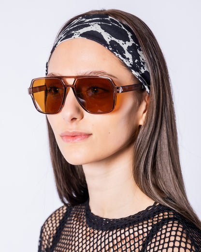 Eine Frau trägt die Urban Aviator Sonnenbrille mit eckigen, braun getönten Gläsern, transparent braunem Rahmen und markantem Doppelsteg. Kombiniert mit einem schwarz-weißen Leo-Stirnband und einem schwarzen Netzoberteil wird die Brille in einer Nahaufnahme vor hellem Hintergrund präsentiert.
