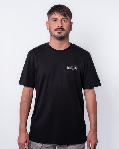 (DE) Ein Mann trägt ein schwarzes T-Shirt mit dem weißen 10 Years PAROOKAVILLE Schriftzug auf der rechten Oberseite. (EN) A man is wearing a black T-shirt with the white 10 Years PAROOKAVILLE lettering on the top right.