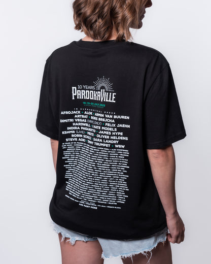 (DE) Eine Frau trägt das schwarze Line-Up T-Shirt.  Auf dem Backprint steht in weißer Schrift das 10 Years PAROOKAVILLE- Schriftemblem und die Künstler. Die Festivaldaten sind grün. (EN) A woman  is wearing the black line-up T-shirt.  The 10 Years PAROOKAVILLE logo and the artists are written in white on the back print. The festival dates are in green.