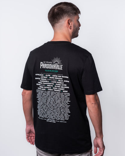(DE) Ein Mann trägt das schwarze Line-Up T-Shirt.  Auf dem Backprint steht in weißer Schrift das 10 Years PAROOKAVILLE- Schriftemblem und die Künstler. Die Festivaldaten sind grün. (EN) A man is wearing the black line-up T-shirt.  The 10 Years PAROOKAVILLE logo and the artists are written in white on the back print. The festival dates are in green.